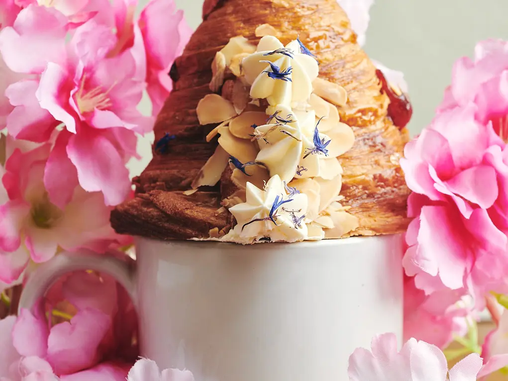 A croissant in a mug surrounded by pink flowers